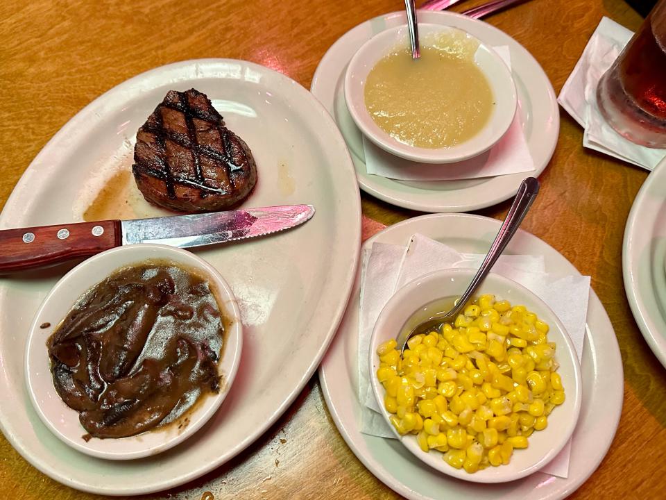 Steak on plate with knife and bowl of mushrooms next to bowl of applesauce and bowl of corn