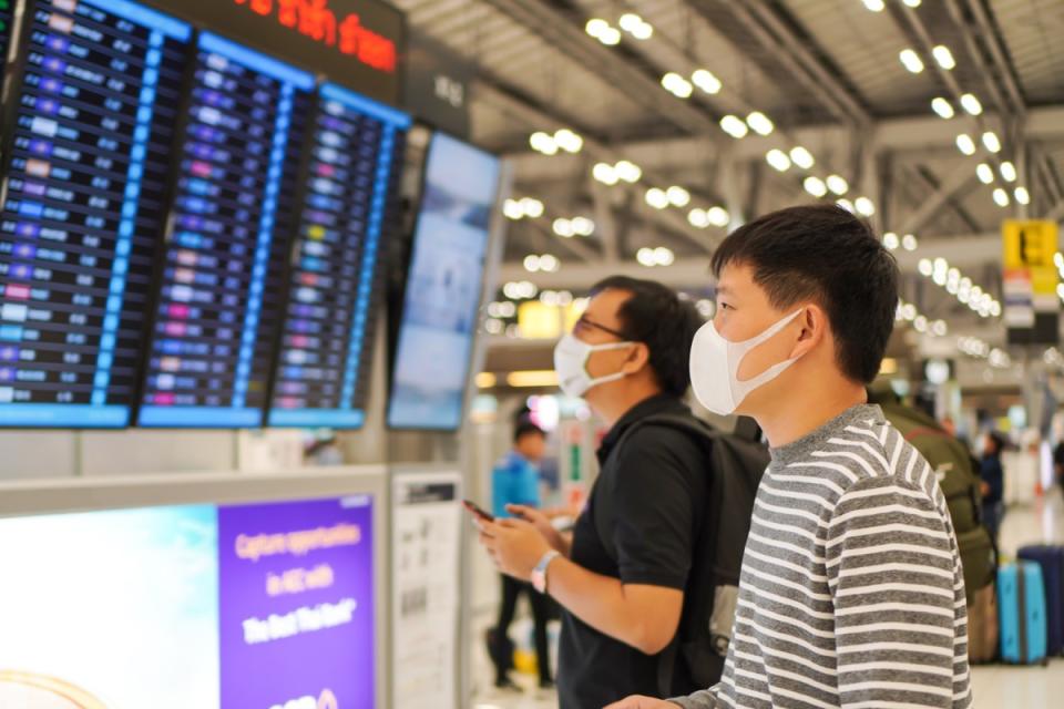 Asian male with face mask protection againts Coronavirus or Covid-19 , Covid19 and omicron  look at flight schedule board at the airport , Corona Virus and health protection concept