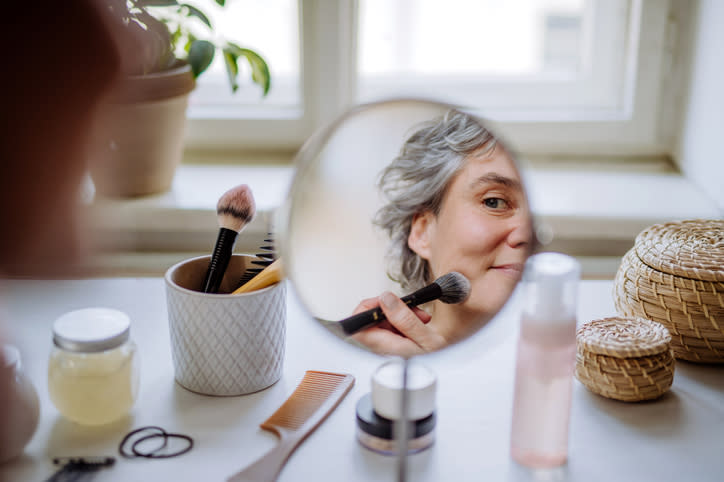 Woman applying makeup while looking into a round mirror, with cosmetic products and brushes nearby