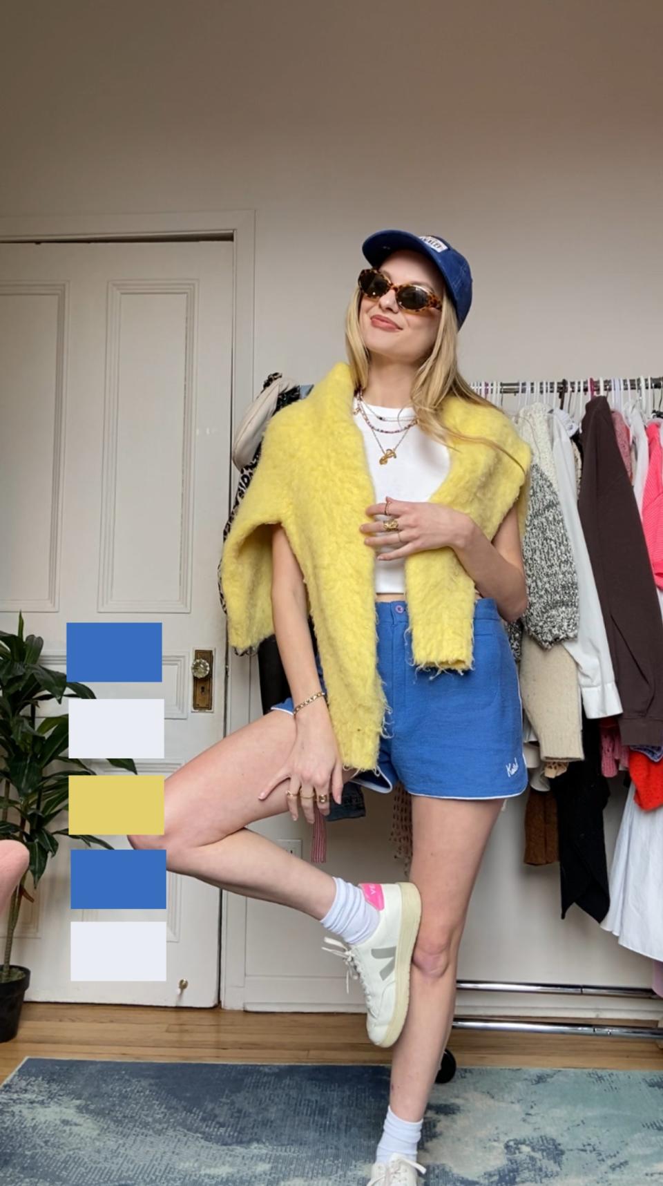 Woman in sunglasses and cap stands posing with one leg raised, wearing a yellow furry jacket and denim shorts