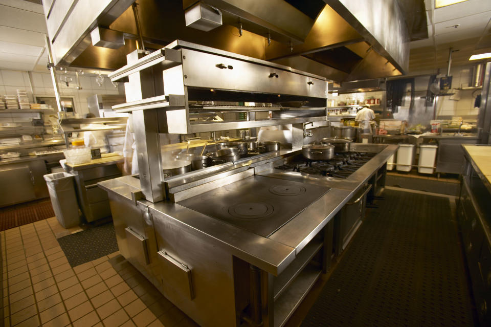 A commercial kitchen with multiple stainless steel appliances and cooking stations. A chef is working in the background