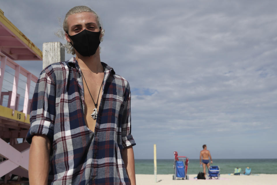 Nivek Divincci wears a protective mask as he poses for a photograph on the beach at Haulover Park during the new coronavirus pandemic, Friday, June 19, 2020, in Miami. Warm weather beach destinations are the most popular vacation searches, with Florida, Myrtle Beach, S.C., San Diego and Key West, Fla., among the top considerations. (AP Photo/Lynne Sladky)