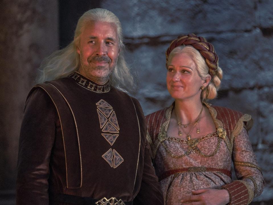 A man and woman with silver hair wearing medieval royalty garb stand lovingly next to each other.