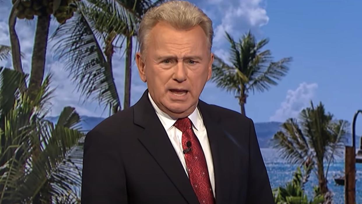  Pat Sajak in black suit and red tie hosting game of Wheel of Fortune. 