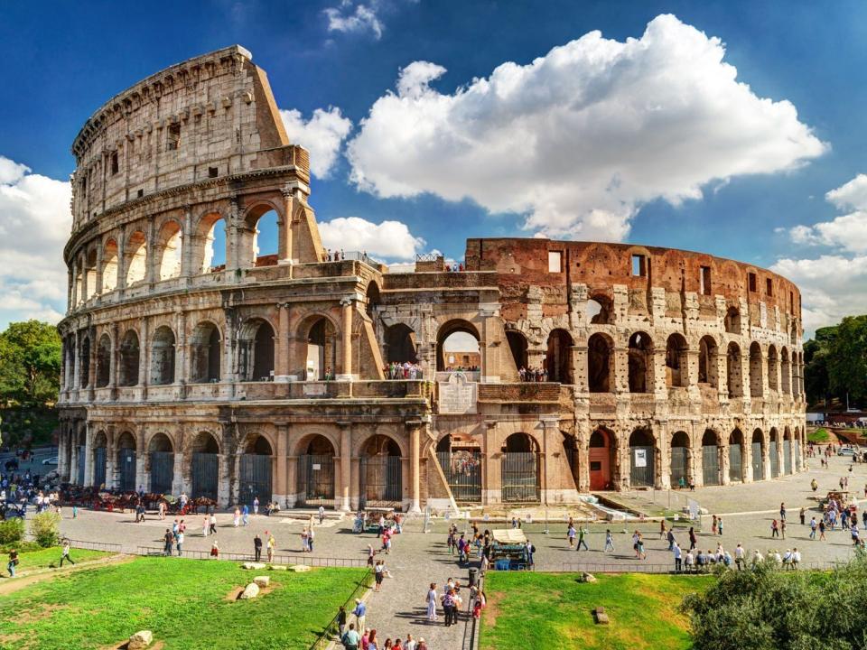 Groups of people walking around the Colosseum and sitting on a large lawn space on a sunny day.