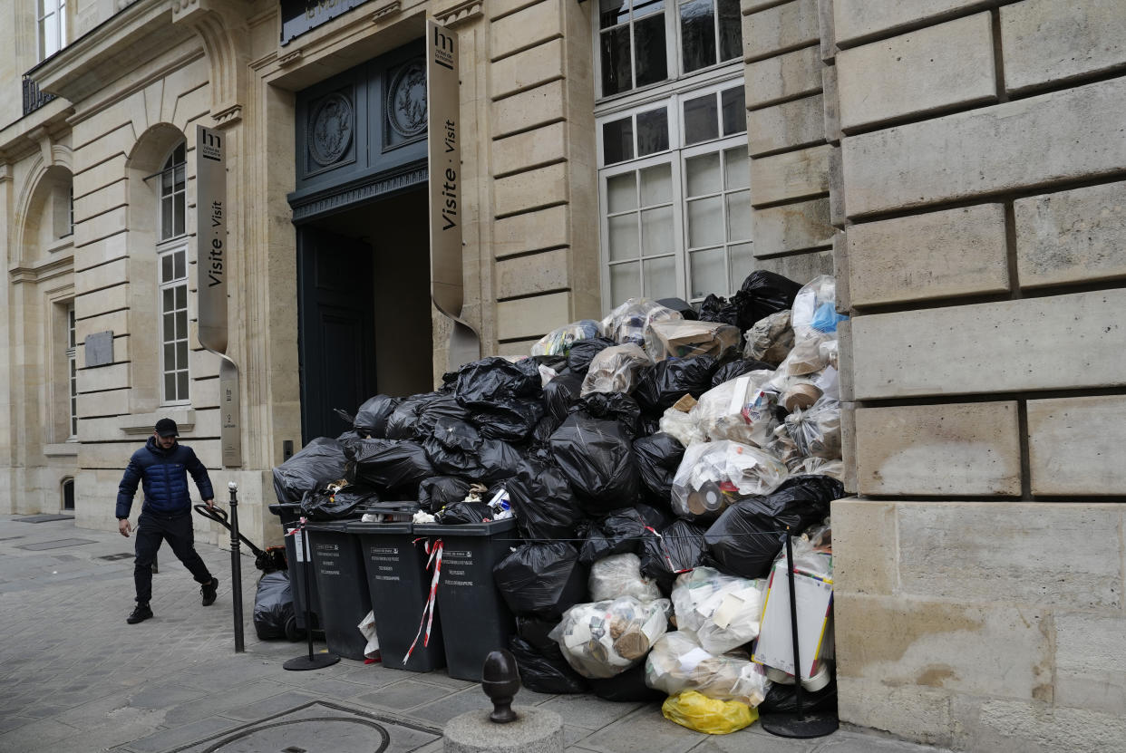 A man walks past uncollected garbages in Paris, Friday March 17, 2023, as sanitation workers are on strike. Protests against French President Emmanuel Macron's decision to force a bill raising the retirement age from 62 to 64 through parliament without a vote disrupted traffic, garbage collection and university campuses in Paris as opponents of the change maintained their resolve to get the government to back down. (AP Photo/Lewis Joly)