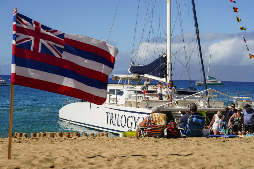 Tourists get off of a Trilogy Excursions boat arriving on Kaanapali Beach in front of a flag of Hawaii planted in the sand, Wednesday, Dec. 6, 2023, in Lahaina, Hawaii. Residents and survivors still dealing with the aftermath of the August wildfires in Lahaina have mixed feelings as tourists begin to return to the west side of Maui. (AP Photo/Lindsey Wasson)