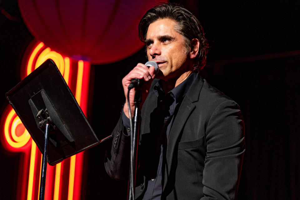 John Stamos in a black suit and dark colored button down shirt speaks into a mic onstage in front of a red neon light and red paper lantern. (Mathieu Bitton / Netflix)