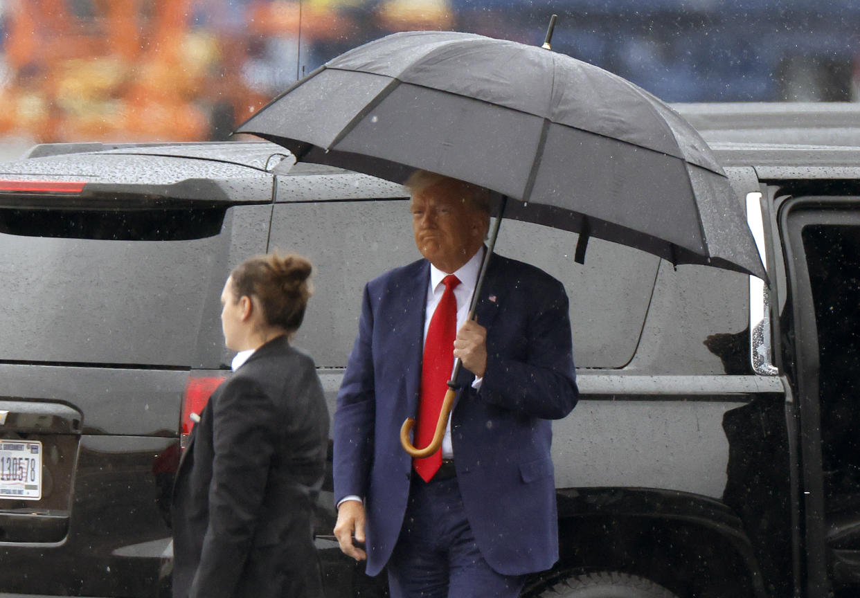 Under an umbrella, Donald Trump, in navy suit and red tie, looks glum.