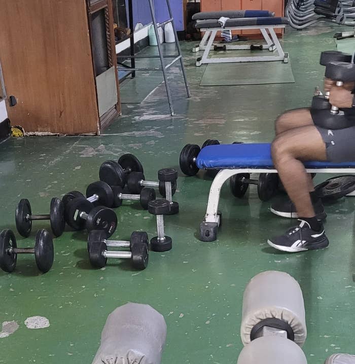 A person is sitting on a bench in a gym, holding a dumbbell in each hand, with several more dumbbells scattered on the floor around them