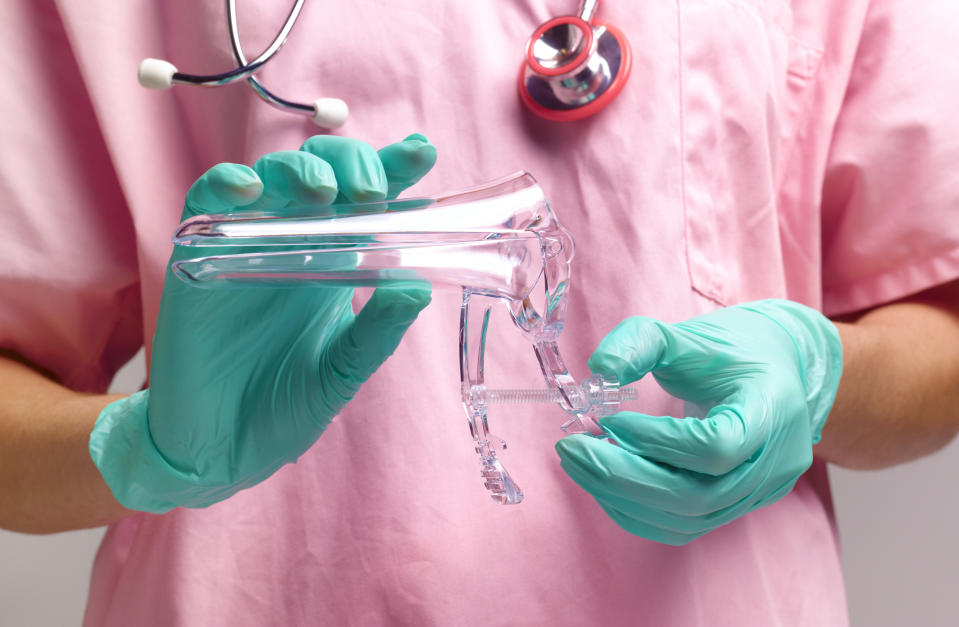 A healthcare professional in pink scrubs and green gloves holds a plastic gynecological speculum and a medical tube