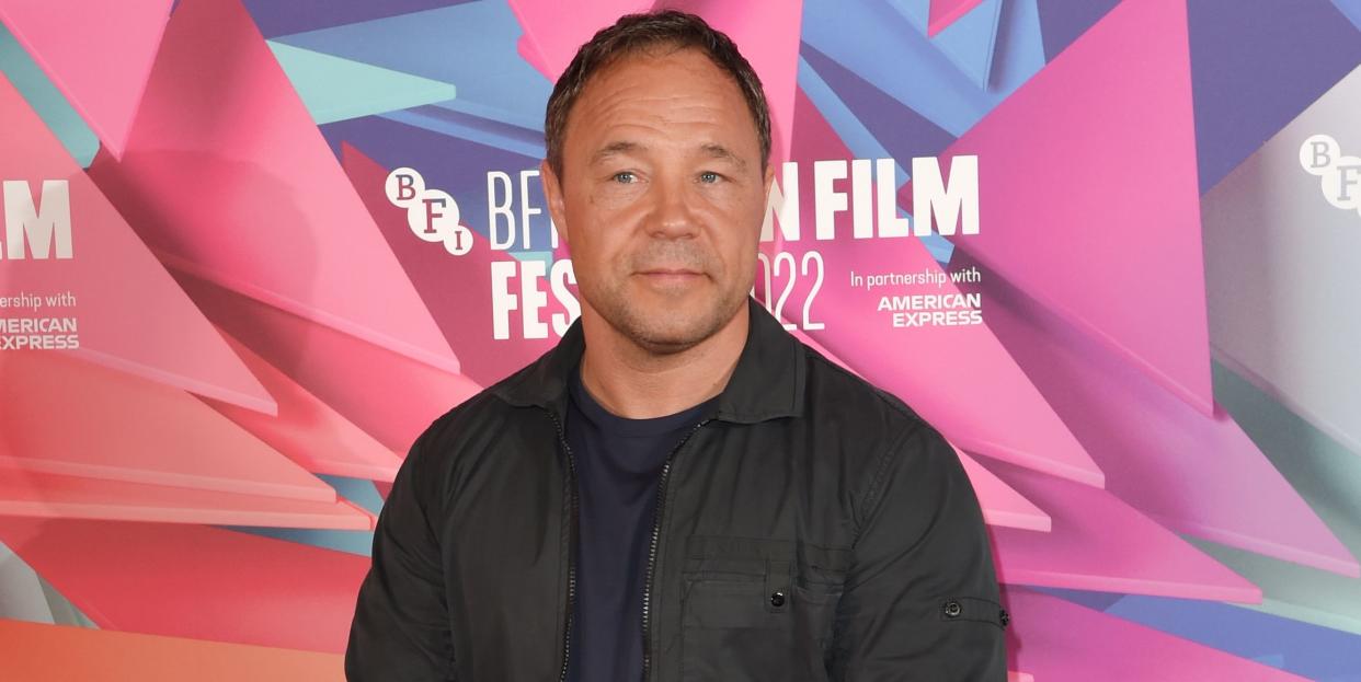 stephen graham, a man stands clasping hands and looking at the camera, he wears black trousers, tshirt, jacket and trainers