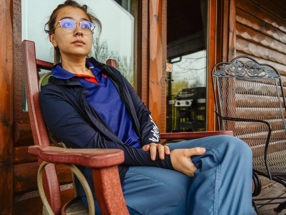 The author sits in a wooden rocking chair in a blue outfit with her legs crossed. Behind her is the cabin with floor-to-ceiling windows. There's another chair on the right.