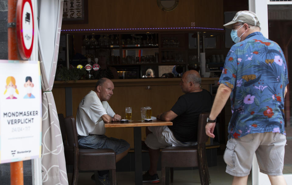 A sign, left, advises people that wearing a protective face mask is obligatory on the street, as two men have a beer in an open cafe at the Belgian seaside resort of Blankenberge, Belgium, Tuesday, Aug. 11, 2020. A skirmish took place on the beach on Saturday, Aug. 8, 2020 which resulted in two coastal communities banning day trippers from the city. (AP Photo/Virginia Mayo)