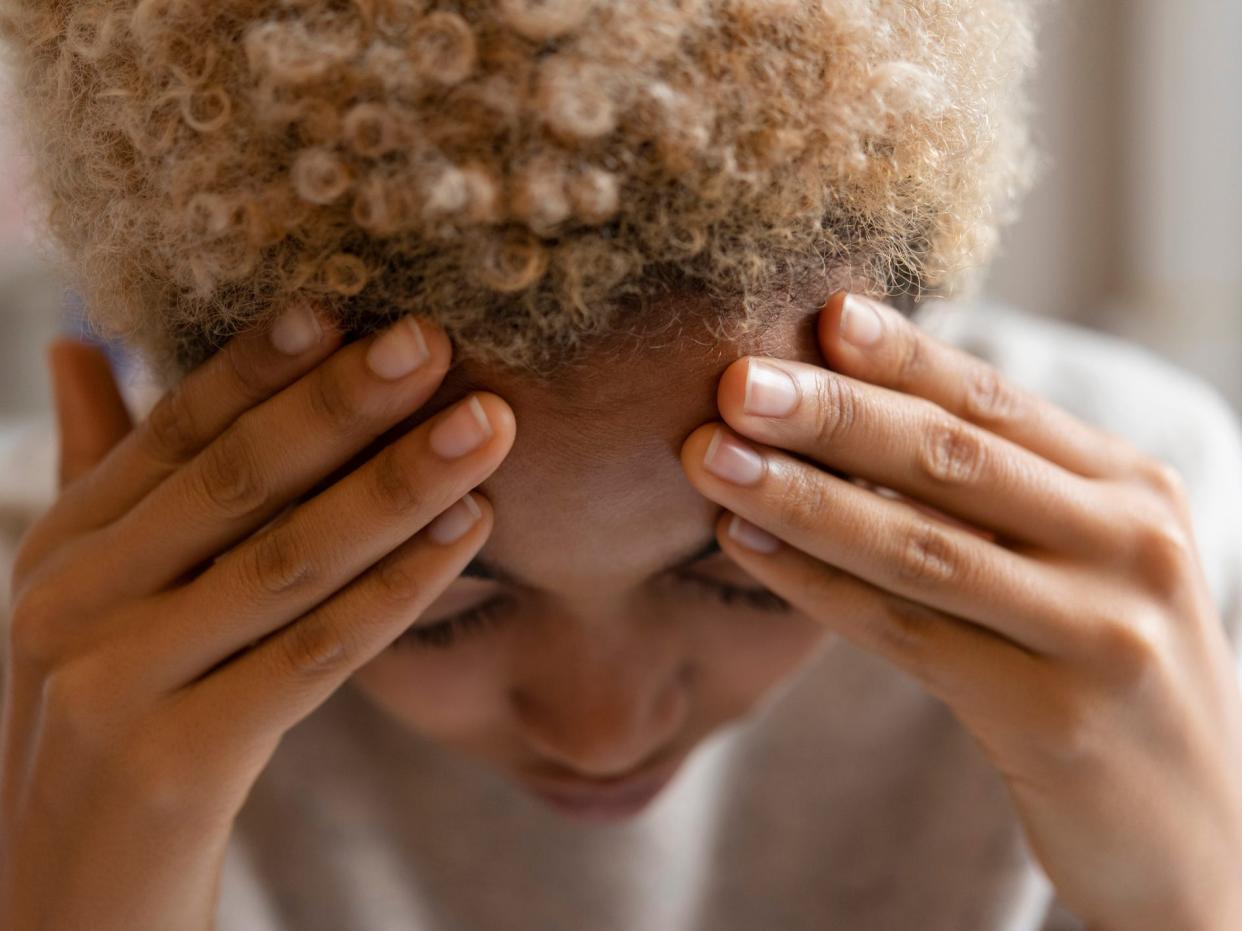 Close-up image of a woman with her face angled downwards, eyes closed, with her hands on her forehead. She has black curly hair in a short Afro, dyed blonde. She wears a tan top.