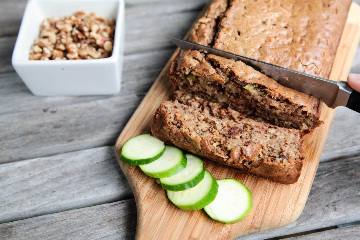Knife cutting through loaf of zucchini bread on a grey wooden table with a white bowl of walnuts in the background