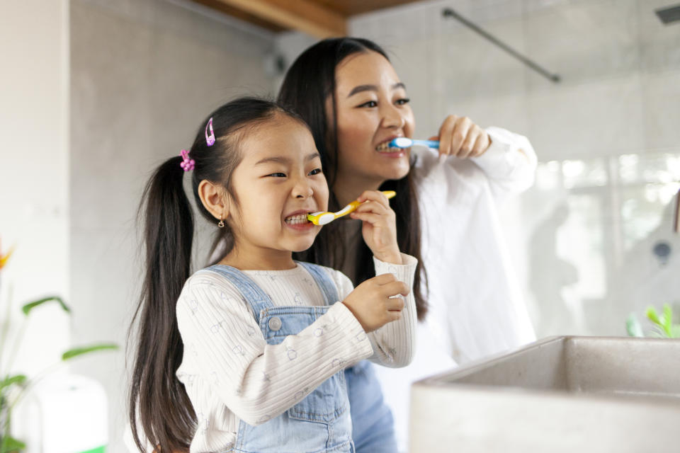 An adult and a child brush their teeth together in a bathroom, smiling into a mirror. The child wears overalls and a long-sleeved shirt