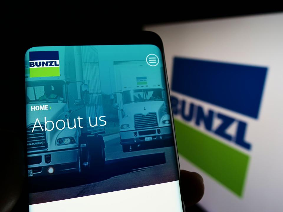 Person holding smartphone with website of British distribution company Bunzl plc on screen in front of logo. Focus on center of phone display.