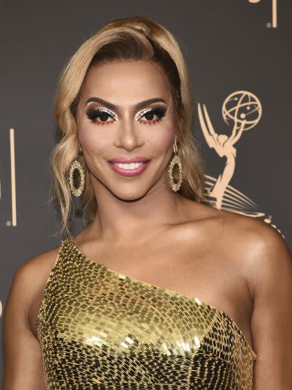 A person with light blonde and brown hair wearing a gold one-shoulder dress smiles