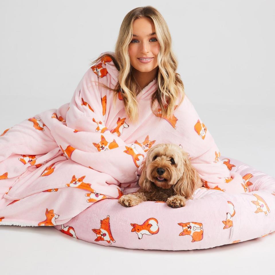 Woman wears an Oodie corgi patterned gown while a tan fluffy dog sits on a same-patterned Oodie Corgi Calming Pet Blanket, $104