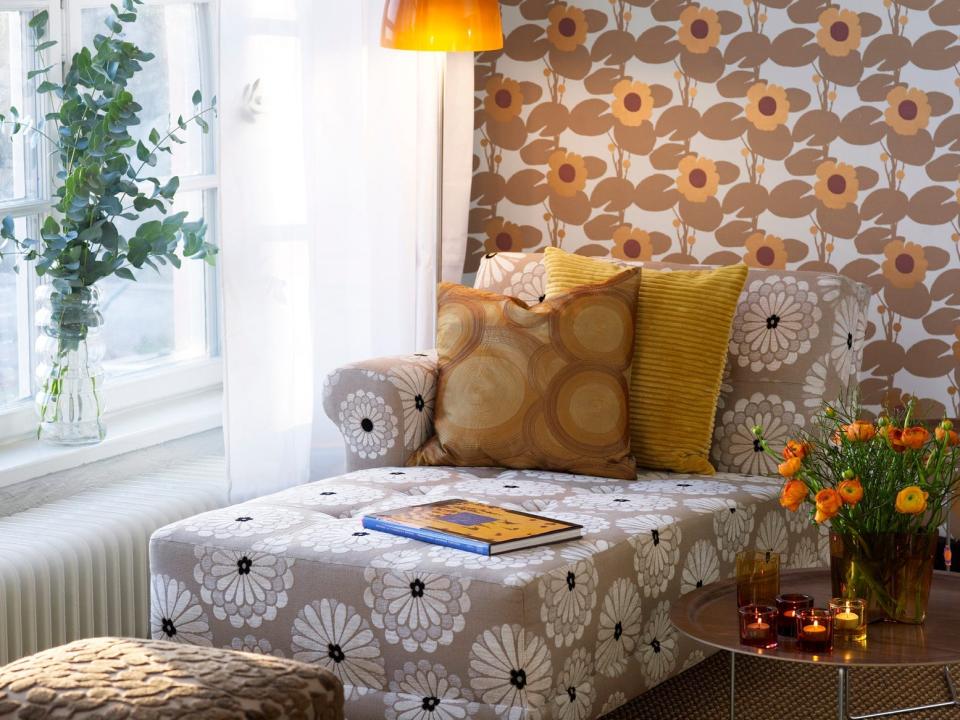 A lounge chair with floral pattern, orange pillows with a circular pattern, a funky floor lamp with an orange shade, and floral wallpaper