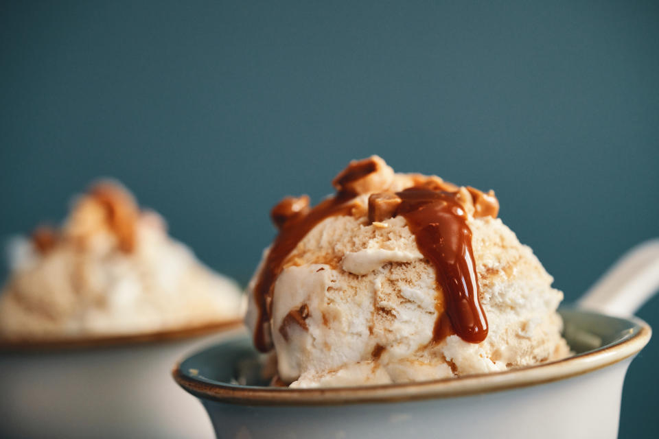 Close-up of a bowl of vanilla ice cream drizzled with caramel sauce and topped with chopped nuts, with another similar bowl in the background