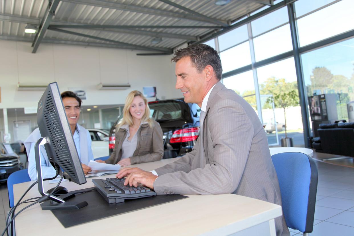 Male car dealer on desktop computer at desk with young couple in car dealership showroom in the background