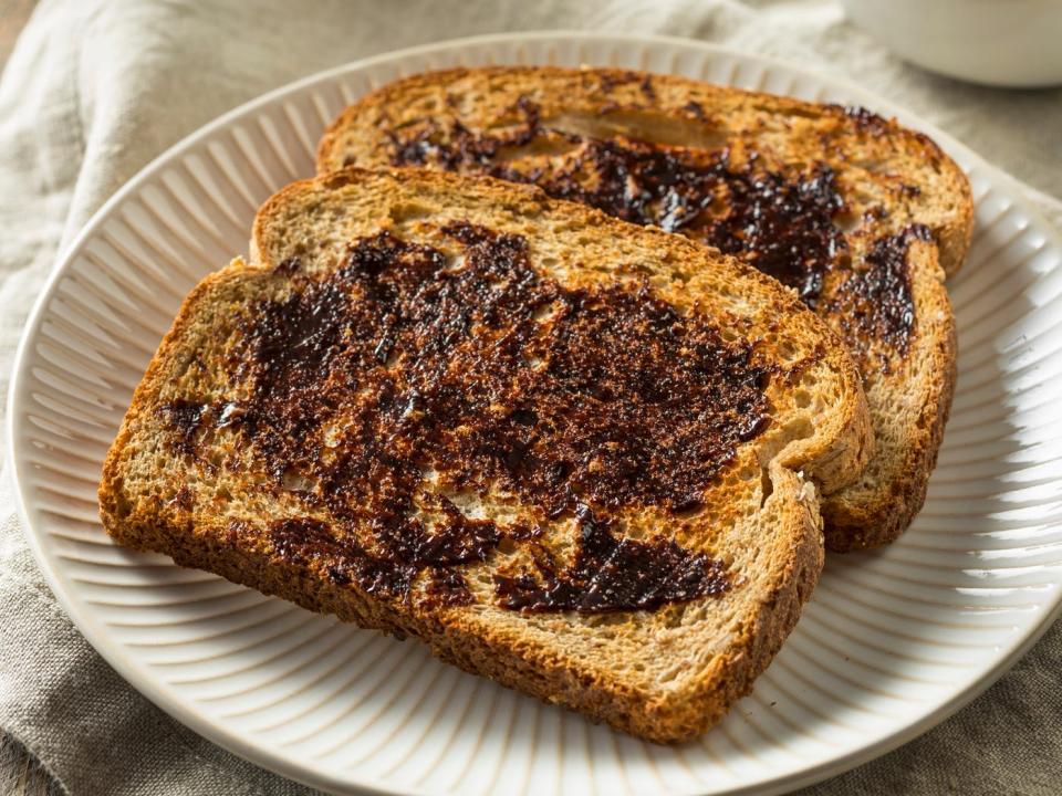 Toast topped with Vegemite on a white plate. There is also a bowl of the spread, a knife, and a tablecloth on the table.