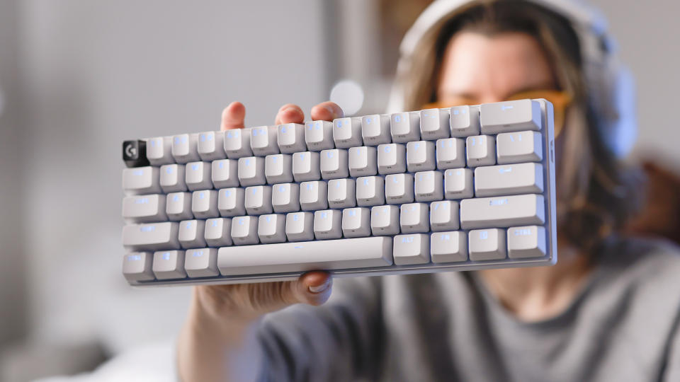 A woman in a gray sweater ،lds up a white Lo،ech G Pro X 60 gaming keyboard up to the camera with her left hand.