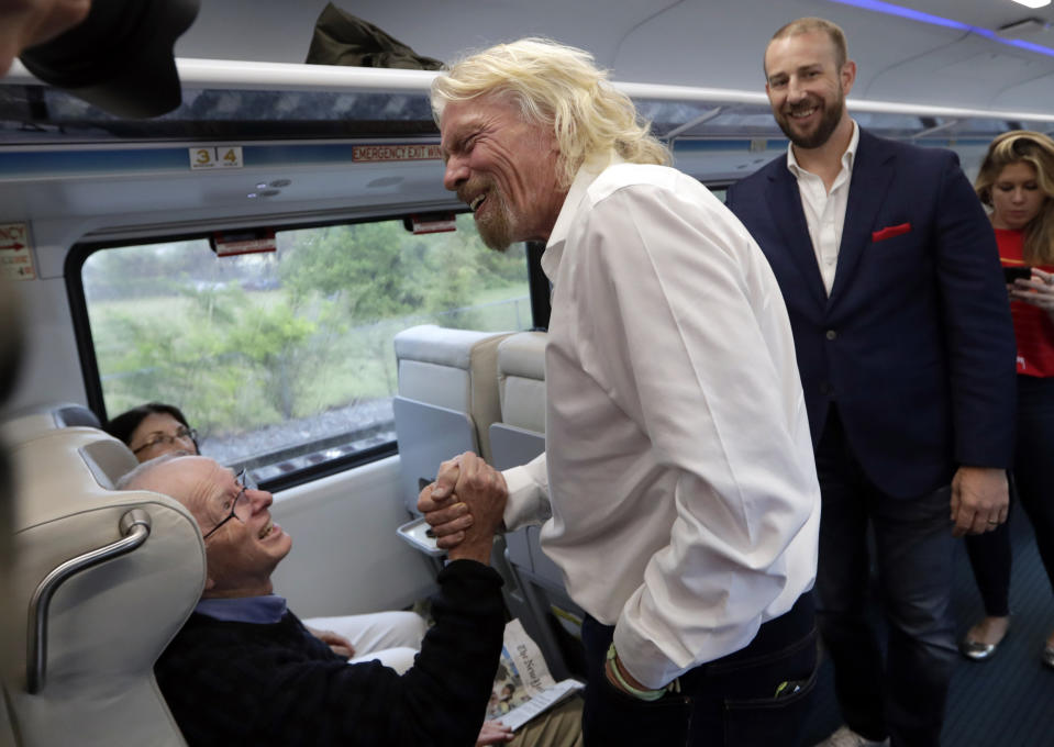 Richard Branson, of Virgin Group, center, greets a passenger while riding a Brightline train from Miami to West Palm Beach, Fla. Thursday, April 4, 2019. The state's Brightline passenger trains are being renamed Virgin Trains USA after Branson invested in the new fast-rail project that is scheduled to connect Miami with Orlando. At right is Patrick Goddard, president of Virgin Trains USA. (AP Photo/Lynne Sladky)