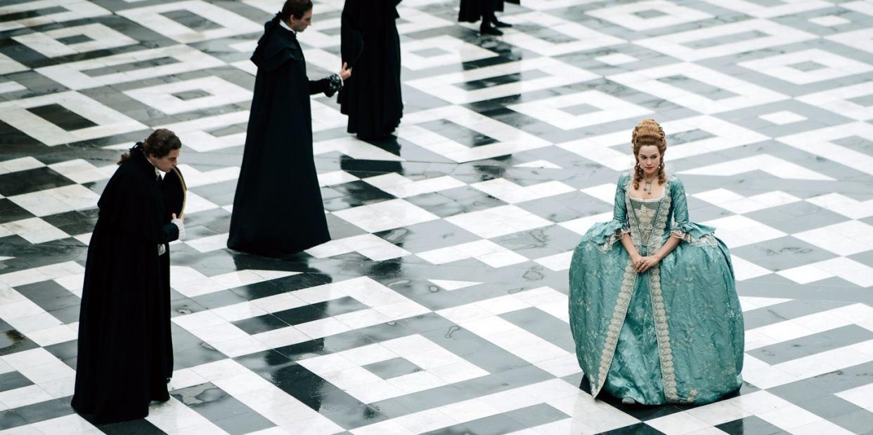 a publicity photo of emilia schule as marie antoinette, on a black and white tiled floor, with four men in black standing behind her