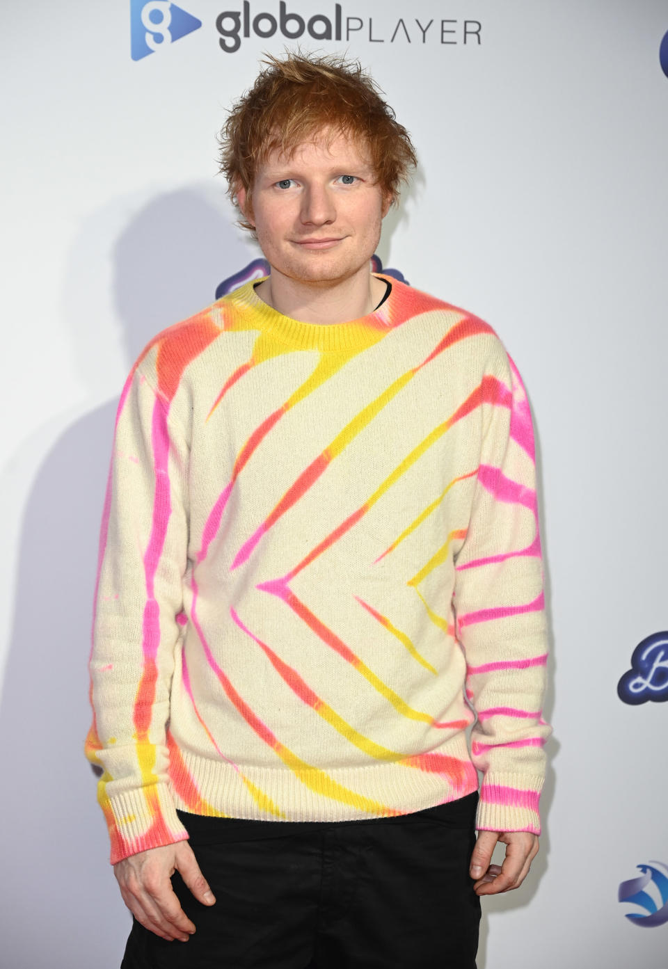 Ed Sheeran at a celebrity event, wearing a sweater with bright, diagonal stripes and black pants