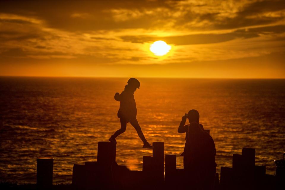 A child climbing on boardwalk posts is silhouetted by a dramatic sunset.