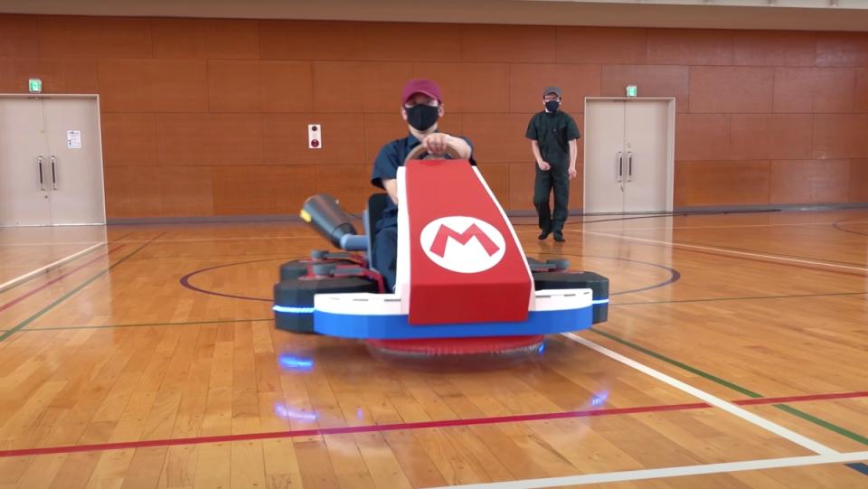 A master cardboard craftsperson driving a real-life hover Mario Kart through a gym while his companion watches him.