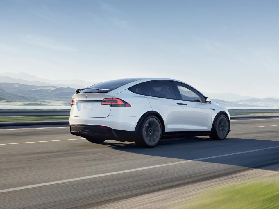 A white Tesla Model X drives down the highway, with blue skies in the background.