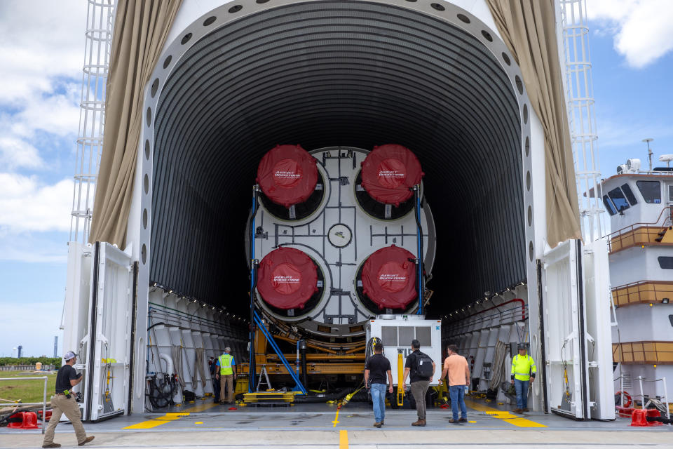 a big rocket is visible from the back, with engines wrapped in plastic. it lies inside a horizontal storage facility. in front, people walk towards the rocket