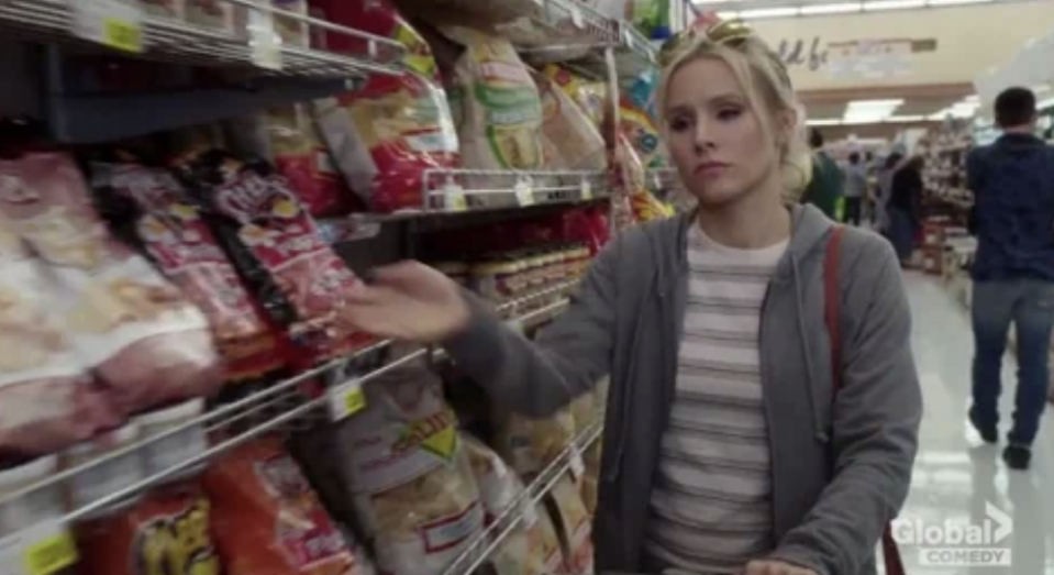 A woman stands in a grocery store snack aisle, gesturing toward a shelf of chips. The scene has a humorous tone