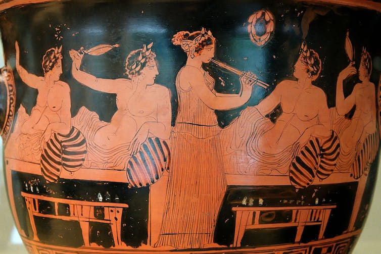 Pottery depicting a woman playing flute-like instrument while topless men recline and listen.