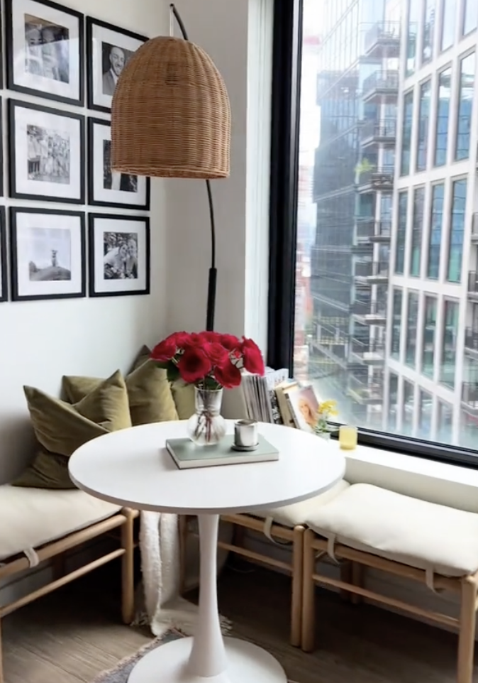 A cozy corner with a small round table, two benches, a floor lamp, and wall frames beside a window overlooking buildings