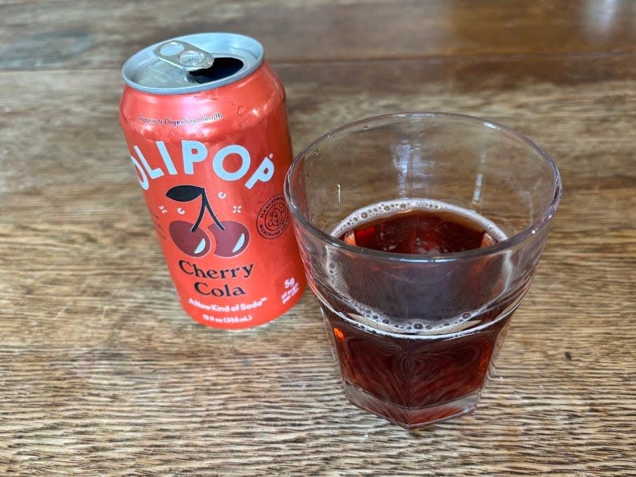 An open can of cherry-cola Olipop next to a small, clear glass with brown liquid inside. Both are sitting on a wooden table.