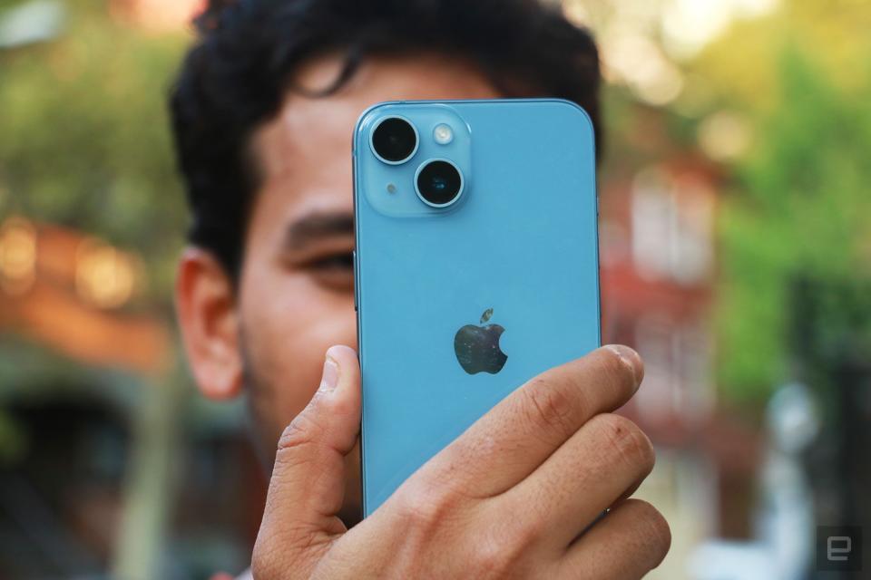 The iPhone 14 held up in mid-air with a man behind it, showing the dual rear cameras.