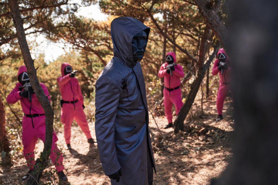 Masked guards in pink suits with triangle symbols holding guns and a masked man in a black hooded coat stand in a forest scene from the show "Squid Game"