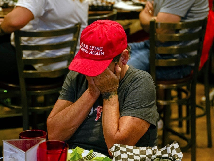 A woman wearing a green shirt and a red hat that reads "Make Lying Young Again" buries her face in her hands at a dinner table.
