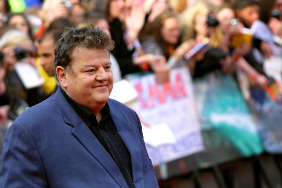 A man wearing a blue jacket and a dark shirt smiles while standing in front of a barricaded crowd