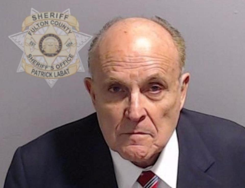 A police mugshot showing Rudy Giuliani in a blue suit and red and blue striped tie which appears to be based on the Stars and Stripes