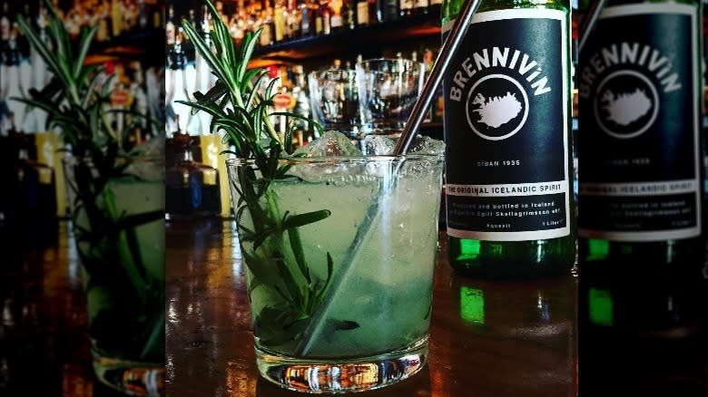 A rosemary-garnished cocktail next to bottle of brennivín