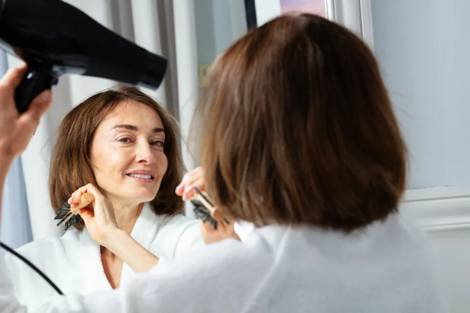 Close-up beauty portrait of a beautiful middle aged woman in housecoat holding hairdryer dry hair before makeup mirror