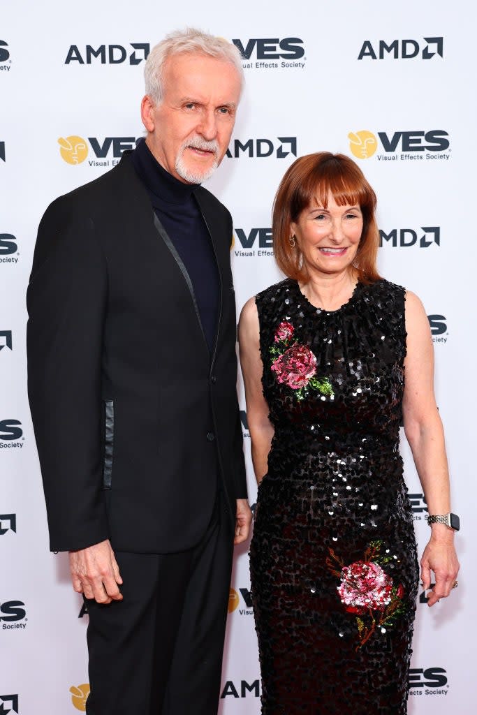 Two individuals posing together, one in a black suit and the other in a sequined dress with floral accents