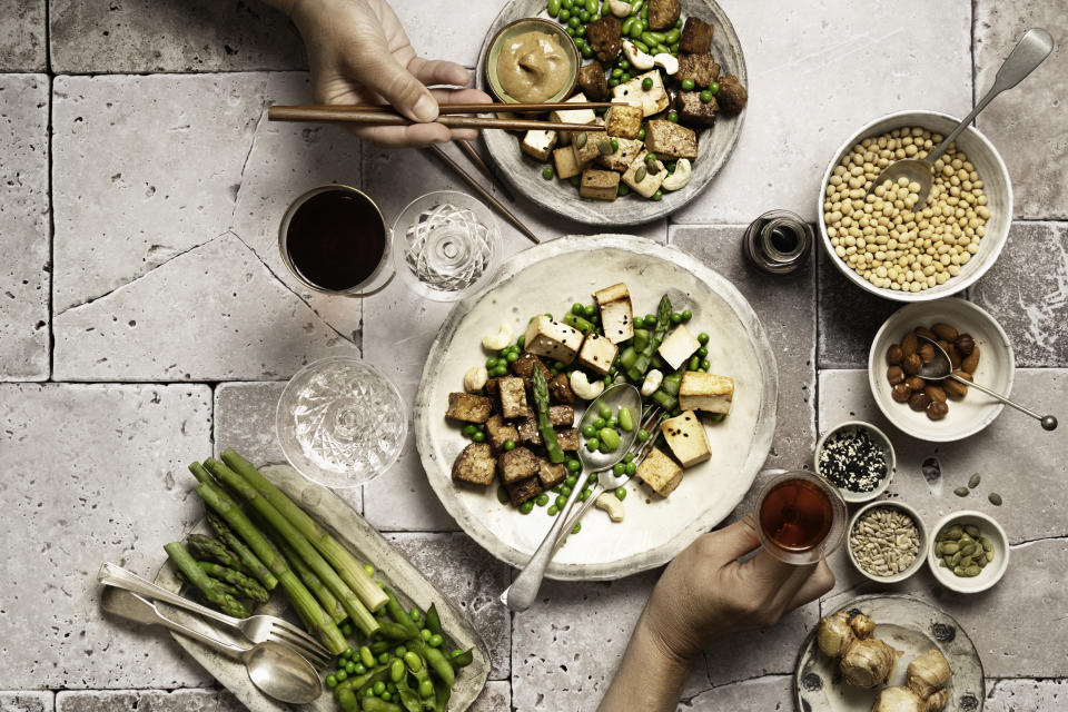 Directly above, a picture of vegan and vegetarian takeaway meals on plates and in bowls with human hands holding glasses and various bowls of plant-based protein sources, miso sauce, seeds, asparagus, edamame and peas. Space to copy