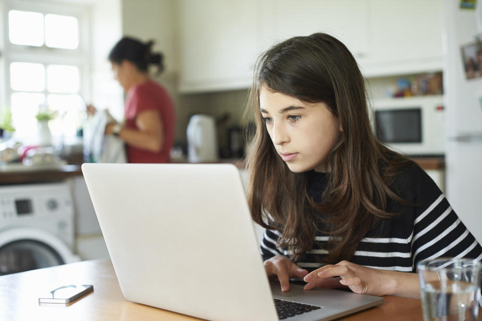 Tween girl on a laptop at home in the kitchen while her mother is at the sink in the background